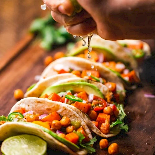 Tacos garnished with vegetables and chickpeas, topped with lime juice being squeezed over them on a wooden board.