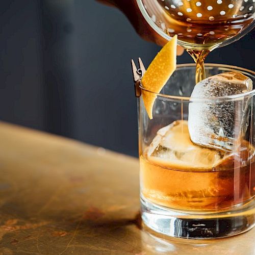 A drink is being poured into a glass with ice and a citrus peel garnish, using a strainer.