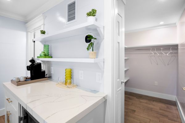 A small kitchen counter with a coffee maker, cups, and greenery; next to it is an open closet with empty hangers and shelves.