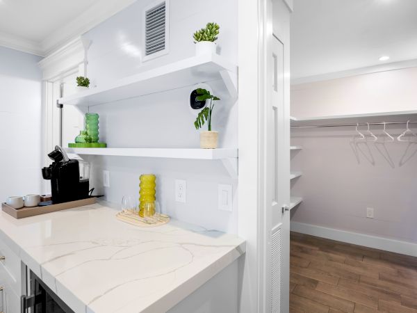 A small kitchen counter with a coffee maker, cups, and greenery; next to it is an open closet with empty hangers and shelves.