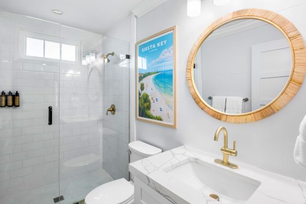 Modern bathroom with gold fixtures, round mirror, white tiles, glass shower, and a "Siesta Key" Florida beach poster on the wall.