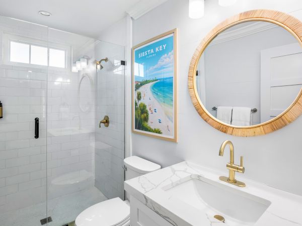 Modern bathroom with gold fixtures, round mirror, white tiles, glass shower, and a "Siesta Key" Florida beach poster on the wall.