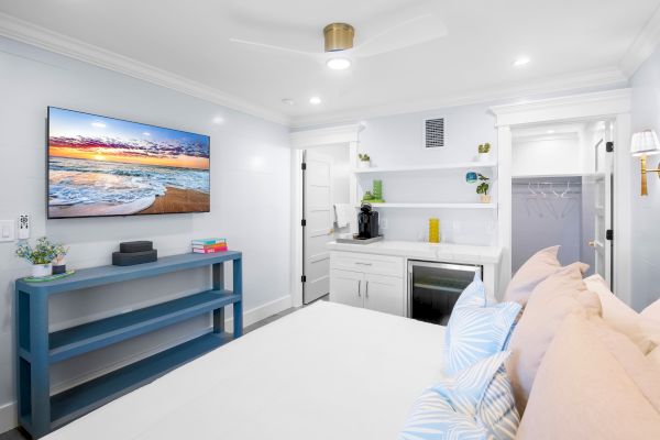 A cozy bedroom with a bed, pillows, wall-mounted TV, blue console, shelving, mini-fridge, and closet in a bright minimalist design.