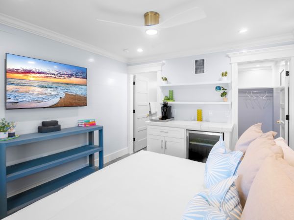 A cozy bedroom with a bed, pillows, wall-mounted TV, blue console, shelving, mini-fridge, and closet in a bright minimalist design.
