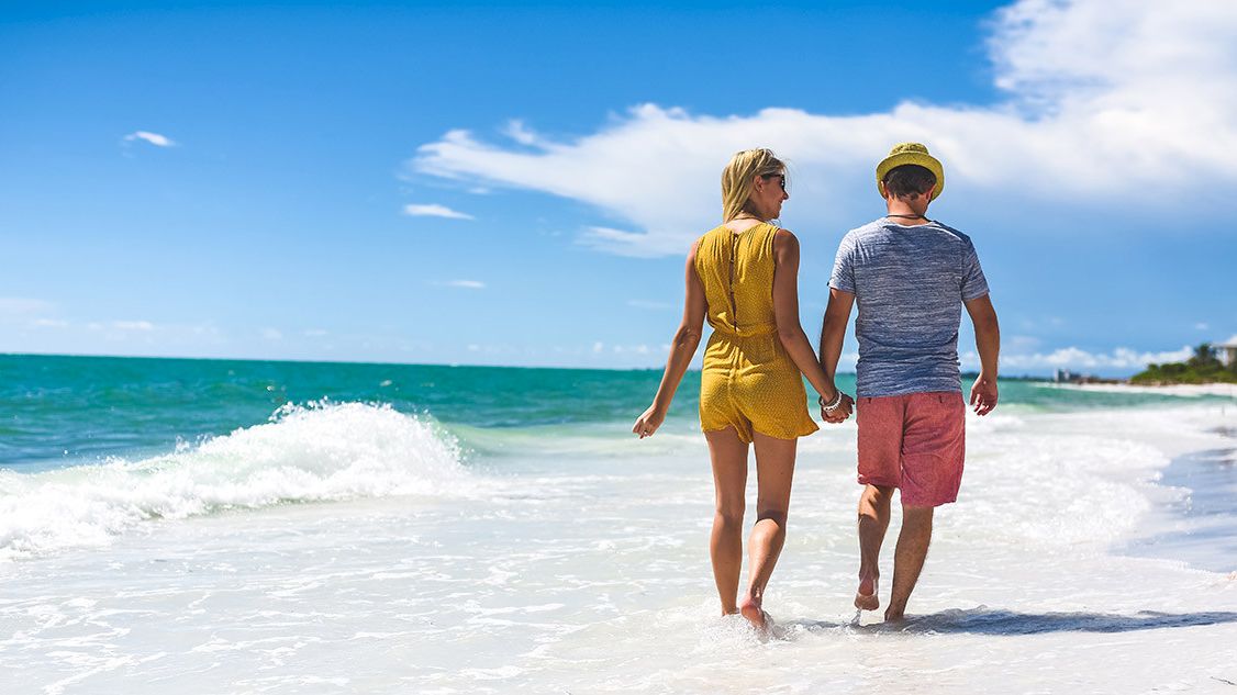 A couple is walking hand in hand along a sunny beach with waves gently lapping at their feet.
