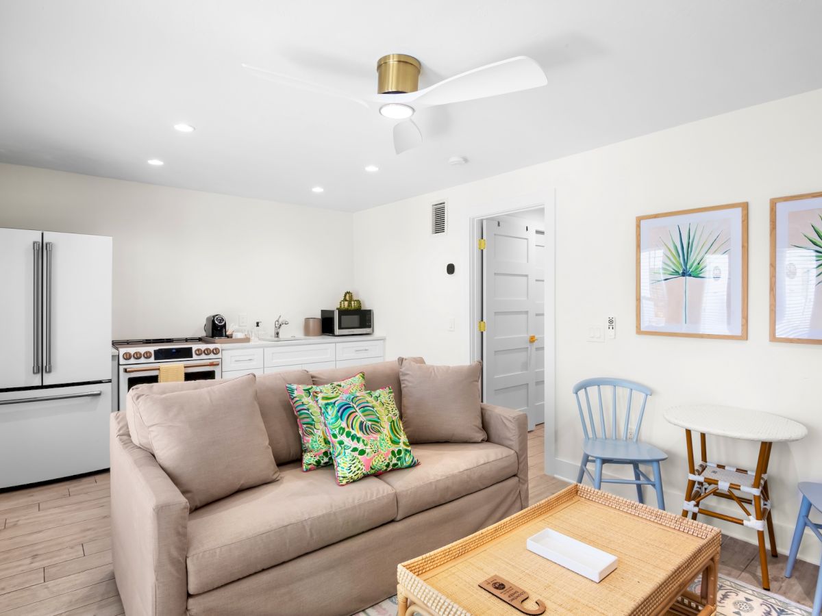 A small living room with a sofa, colorful cushions, coffee table, fridge, stove, and artwork on walls, featuring a modern ceiling fan.