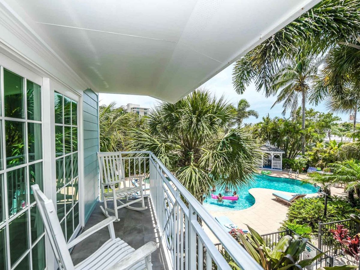 A balcony with white rocking chairs overlooks a tropical pool area surrounded by palm trees and lush greenery.