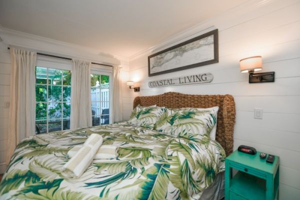 A cozy bedroom features tropical bedding, a woven headboard, coastal decor, and a teal nightstand with a clock, next to a window.