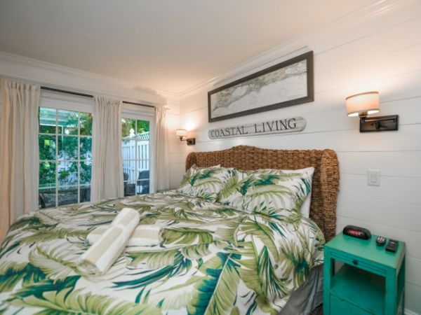 A cozy bedroom features tropical bedding, a woven headboard, coastal decor, and a teal nightstand with a clock, next to a window.