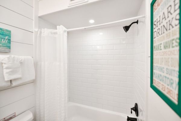 The image shows a bright bathroom with white tile walls, a shower curtain, towels, and a decorative sign on the wall.