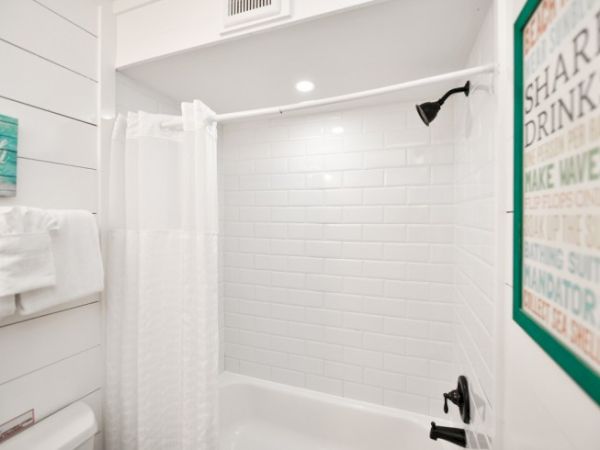 The image shows a bright bathroom with white tile walls, a shower curtain, towels, and a decorative sign on the wall.