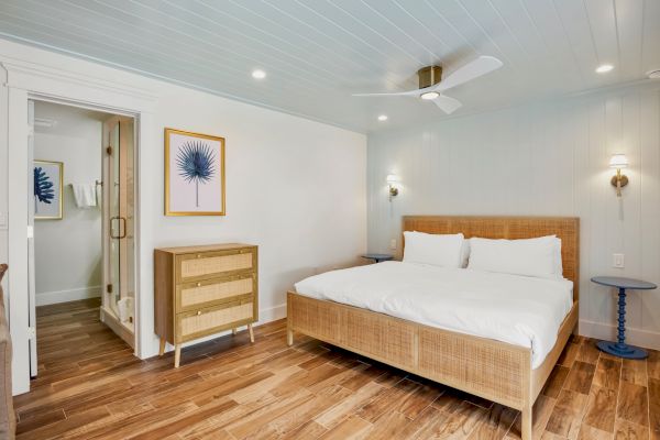 A bedroom with a wooden bed, white bedding, two blue side tables, a dresser, wall art, and a fan. There's an attached bathroom visible.