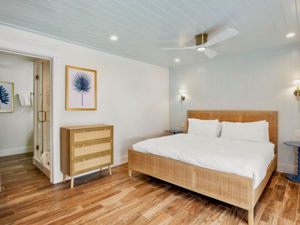 A bedroom with a wooden bed, white bedding, two blue side tables, a dresser, wall art, and a fan. There's an attached bathroom visible.