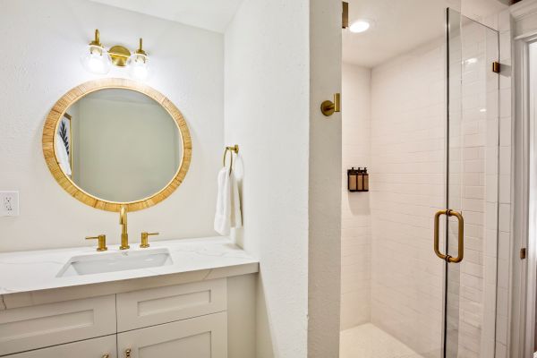 A bathroom with a round mirror, gold fixtures, white vanity, glass shower door, and towel holder on a white wall.