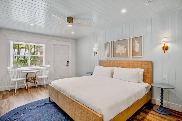 A bright bedroom with a wooden bed, white linens, small table, chairs by the window, and wall-mounted lights on each side of the headboard.