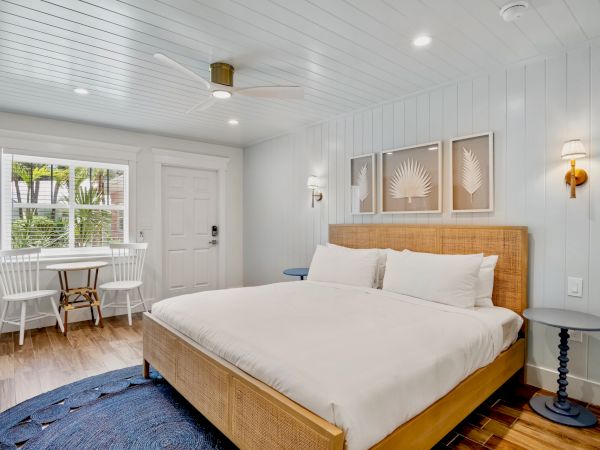 A bright bedroom with a wooden bed, white linens, small table, chairs by the window, and wall-mounted lights on each side of the headboard.