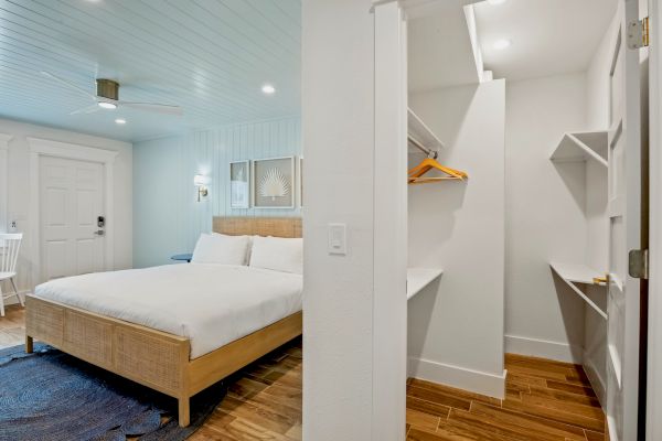 A modern bedroom with a wooden bed, white linens, open closet, wooden floor, and minimal decor is shown.
