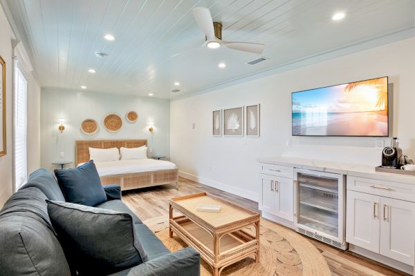 A bright, modern bedroom with a bed, sofa, TV, and kitchenette. Coastal decor and wood accents create a relaxing atmosphere.