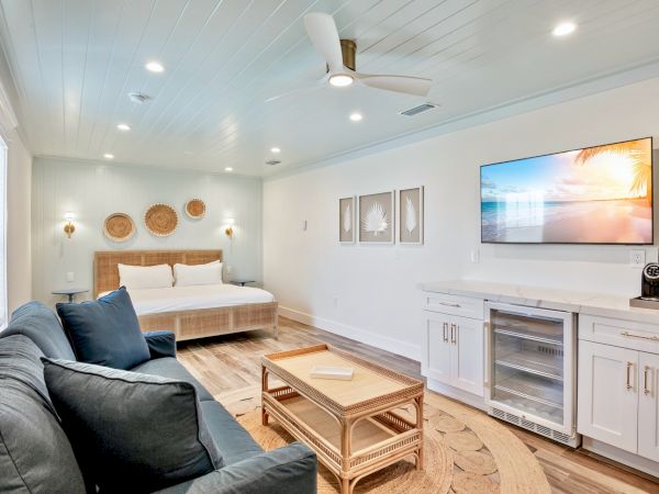 A bright, modern bedroom with a bed, sofa, TV, and kitchenette. Coastal decor and wood accents create a relaxing atmosphere.