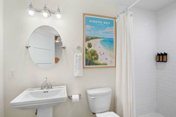 A bathroom with a sink, round mirror, toilet, shower, and Siesta Key poster decorates the wall.