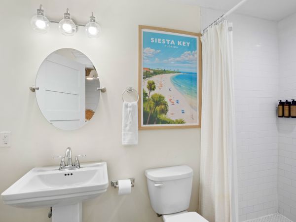 A bathroom with a sink, round mirror, toilet, shower, and Siesta Key poster decorates the wall.