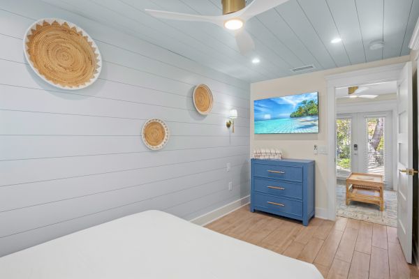 A bedroom with light blue walls, three woven wall hangings, a blue dresser, and a beach scene on TV. A doorway leads outside.