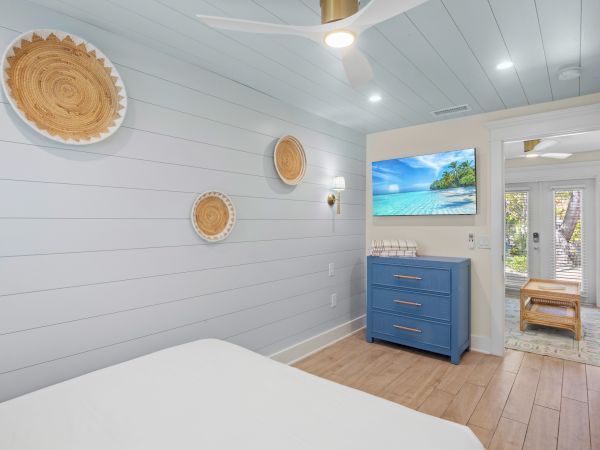 A bedroom with light blue walls, three woven wall hangings, a blue dresser, and a beach scene on TV. A doorway leads outside.