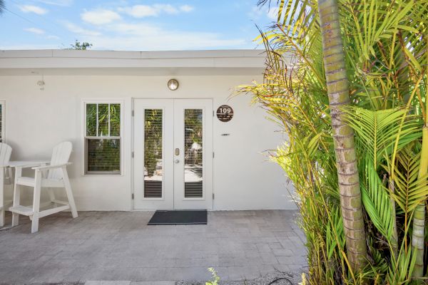 A white building with double glass doors, a chair set, and lush green plants against a clear blue sky.