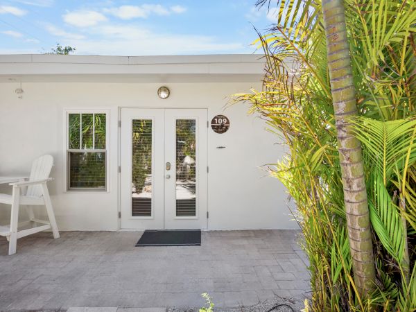 A white building with double glass doors, a chair set, and lush green plants against a clear blue sky.