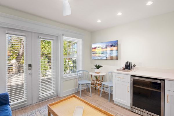 A bright room with glass doors, a small table, chairs, cabinet, coffee maker, and wall art, opening to a patio.