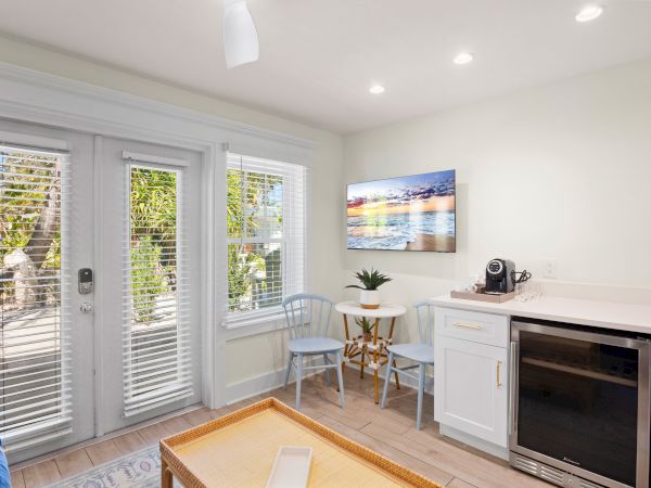 A bright room with glass doors, a small table, chairs, cabinet, coffee maker, and wall art, opening to a patio.