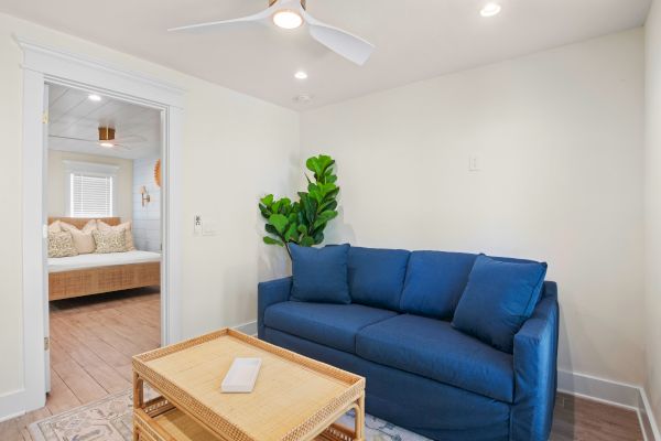 A living room with a blue sofa, a small coffee table, a plant, and a view of a bedroom with a bed in the background.