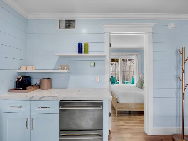 A cozy corner with a countertop, coffee maker, shelves, and entryway to a bedroom, painted in soft pastels.