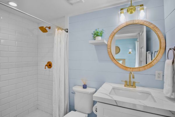 A modern bathroom with a shower, toilet, and sink. Features a round mirror, blue walls, and gold fixtures, creating a stylish look.