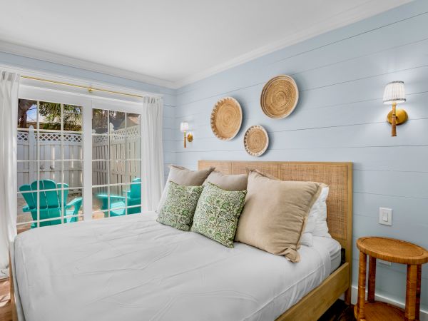 A cozy bedroom with a bed, decorative pillows, woven wall art, wall sconces, and a sliding door leading to a patio with turquoise chairs.