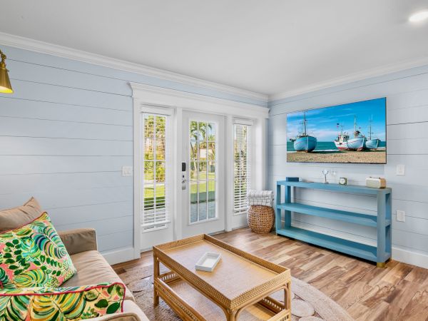 A cozy living room with light blue walls, a sofa with colorful cushions, a wicker coffee table, and a TV displaying boats.