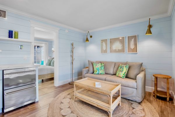 A cozy living room with a beige sofa, colorful pillows, wicker furniture, light blue walls, decorative wall art, and a small fridge.