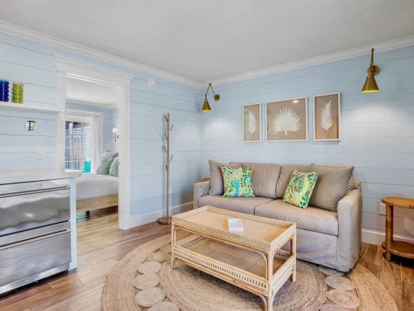 A cozy living room with a beige sofa, colorful pillows, wicker furniture, light blue walls, decorative wall art, and a small fridge.