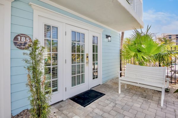 A light blue house entrance with French doors, a door number 185, potted plants, and a white bench on a stone pathway with a palm.