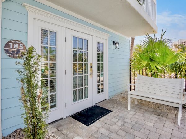 A light blue house entrance with French doors, a door number 185, potted plants, and a white bench on a stone pathway with a palm.