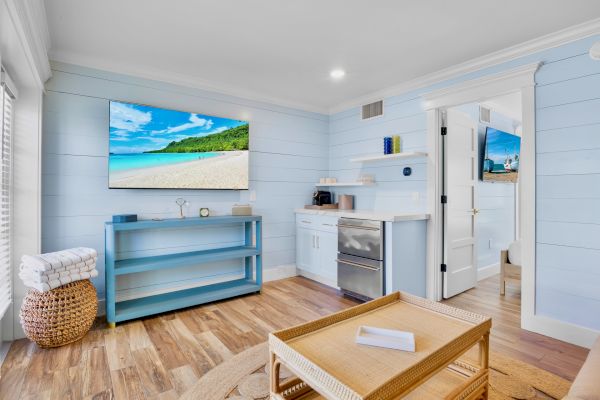 A cozy, bright room with light blue walls, a TV displaying a beach scene, woven furniture, and a small kitchenette area visible.