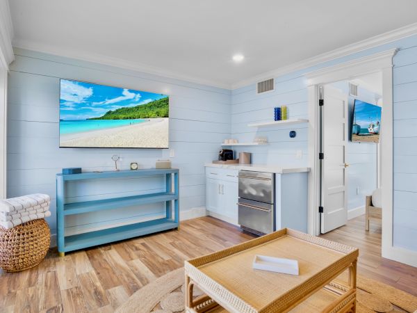 A cozy, bright room with light blue walls, a TV displaying a beach scene, woven furniture, and a small kitchenette area visible.