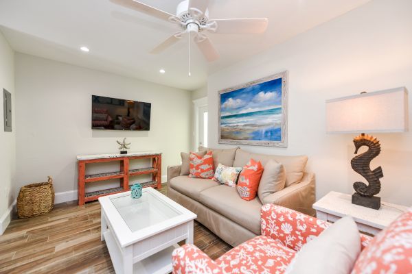 A cozy living room with a sofa, armchair, TV, beach artwork, and seahorse lamp, featuring coastal decor and wooden flooring.