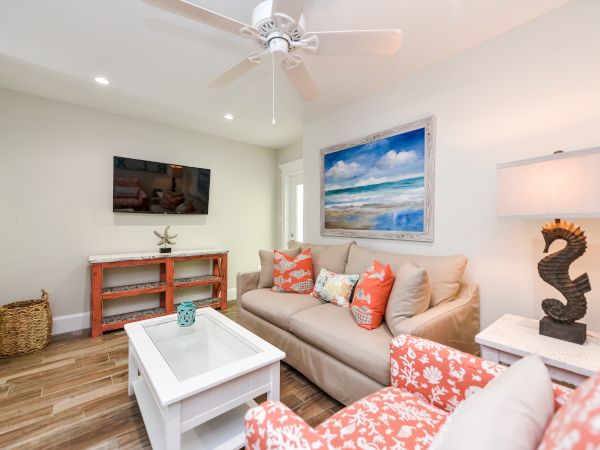 A cozy living room with a sofa, armchair, TV, beach artwork, and seahorse lamp, featuring coastal decor and wooden flooring.
