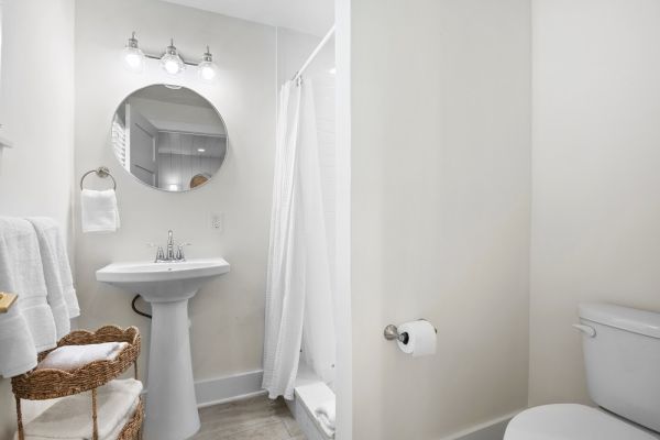 A clean bathroom with a round mirror, pedestal sink, shower with curtain, toilet, towel rack, wicker basket, and overhead lighting.