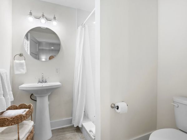 A clean bathroom with a round mirror, pedestal sink, shower with curtain, toilet, towel rack, wicker basket, and overhead lighting.