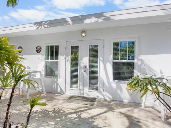 A sunlit patio with white furniture and potted plants, featuring a glass door and windows, marked as room 102 under a clear sky.