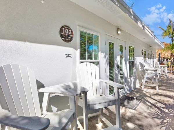 Outdoor seating area with white chairs and tables beside a building. Door marked "102" is visible with shadows of palm trees.