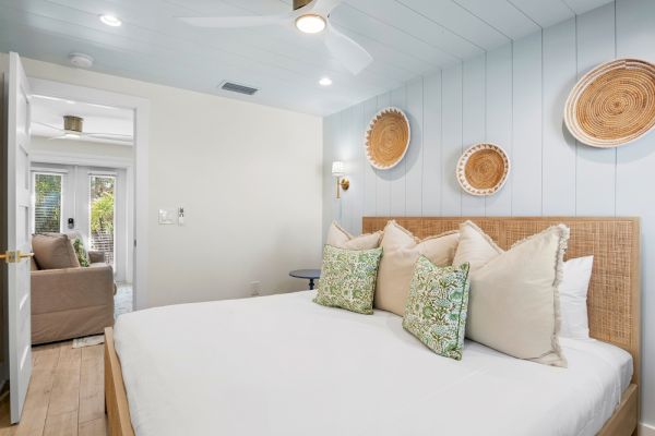 A cozy bedroom with a bed adorned with green and beige pillows, decorative baskets on the wall, and a doorway leading to a sunny room.