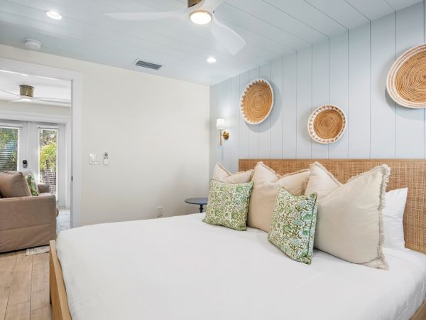 A cozy bedroom with a bed adorned with green and beige pillows, decorative baskets on the wall, and a doorway leading to a sunny room.